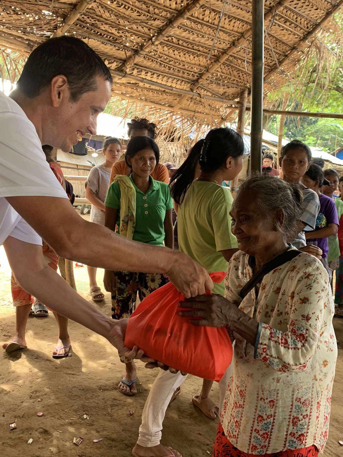 A Children of the Jungle volunteer hands a red bag of food to a smiling Mangyan elder as neighbors look on under a thatched shelter